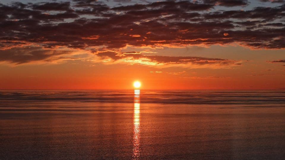 Sun setting into the ocean beneath layered clouds